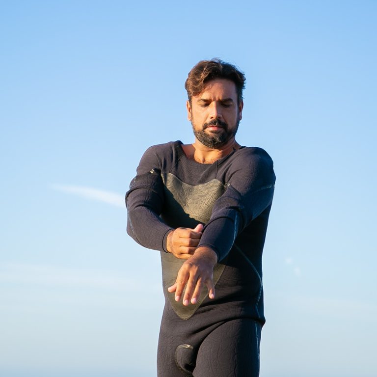 Focused sportsman putting on wetsuit for surfing on ocean beach. Low angle, blue sky background. Active lifestyle concept